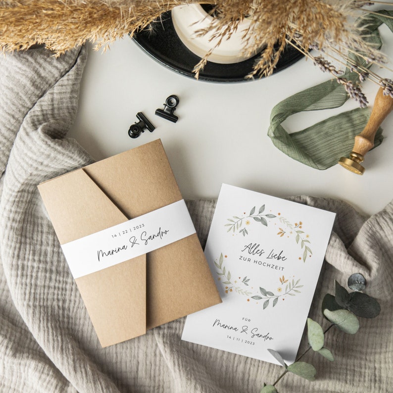 May include: A kraft paper envelope with a white ribbon and the text "14 1 22 1 2023 Marina & Sandro" printed on it. A white card with a floral wreath design and the text "Alles Liebe zur Hochzeit" and "Fur Marina & Sandro 14 1 1 2023" printed on it.