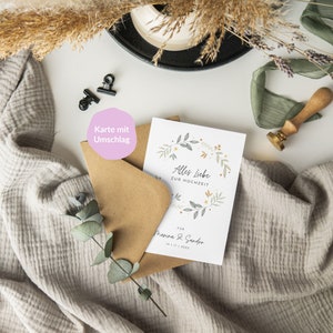 May include: A white card with a floral wreath design and the text "Alles Liebe zur Hochzeit" for Marina & Sandro, 14.11.2023. The card is on a brown envelope with a pink sticker that says "Karte mit Umschlag".