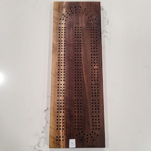 Puede incluir: Un tablero de cribbage de madera con una mancha marrón oscura. El tablero tiene un diseño único con una parte superior curva y una serie de agujeros para clavijas. El tablero está etiquetado con el número "8".