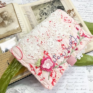 May include: A pink fabric pouch with white lace trim and the words "Sew Happy" embroidered in blue thread. The pouch is decorated with a pink crocheted flower and red floral fabric.