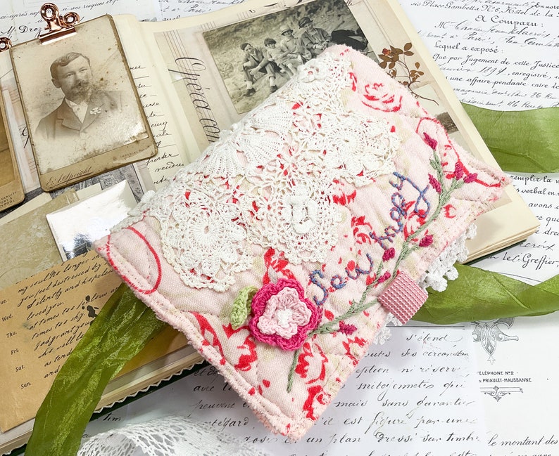 May include: A pink fabric pouch with white lace trim and the words "Sew Happy" embroidered in blue thread. The pouch is decorated with a pink crocheted flower and red floral fabric.