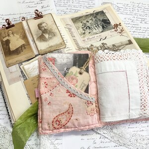 May include: A pink fabric journal with a lace trim and a white handkerchief inside. The journal is decorated with a floral pattern and a paisley design. The journal is surrounded by vintage papers and ephemera.