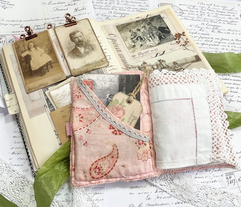 May include: A pink fabric journal with a lace trim and a white handkerchief inside. The journal is decorated with a floral pattern and a paisley design. The journal is surrounded by vintage papers and ephemera.
