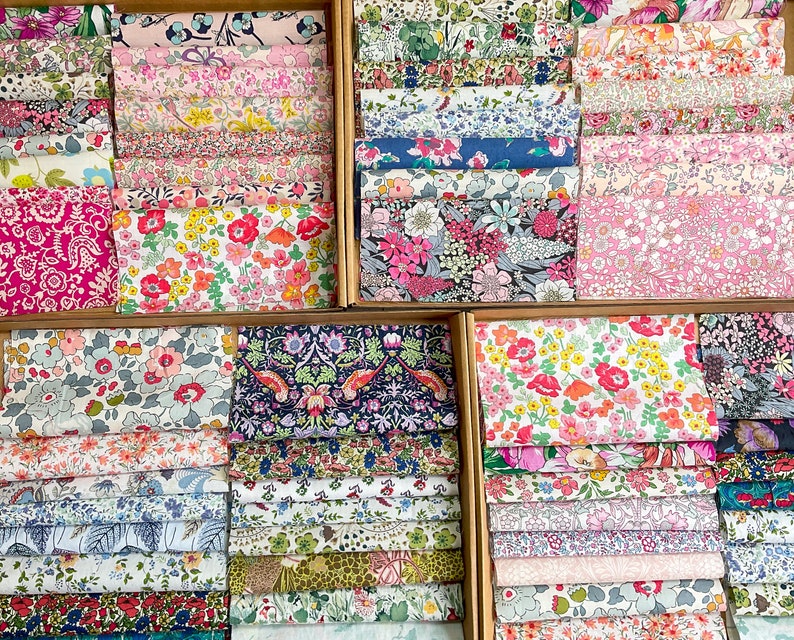 May include: A selection of floral fabric bundles in wooden boxes. The fabrics display a variety of colourful floral patterns, including pink, red, blue, and yellow blooms. The fabrics are neatly folded and stacked, showcasing a range of designs.