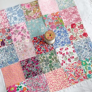 May include: A patchwork quilt made from floral fabric squares in pink, blue, and green hues. A wooden spool of white thread rests on the quilt. The quilt is displayed on a white surface.