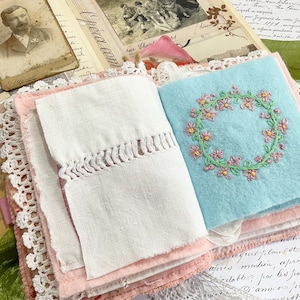 May include: A handmade journal with a pink and white fabric cover. The journal is open to reveal a page with a blue fabric insert featuring a pink embroidered floral wreath. The text "Opera com" is visible on the cover.