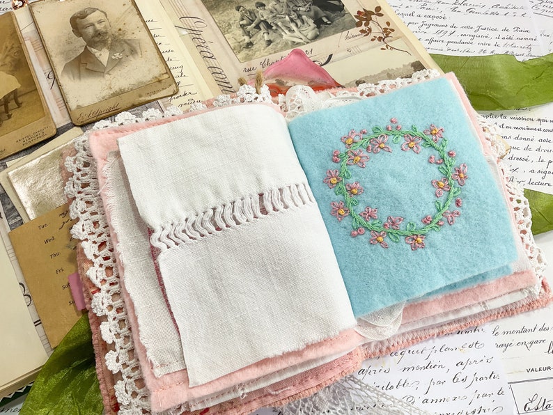 May include: A handmade journal with a pink and white fabric cover. The journal is open to reveal a page with a blue fabric insert featuring a pink embroidered floral wreath. The text "Opera com" is visible on the cover.