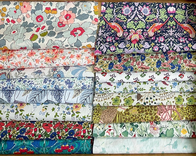 May include: A selection of folded fabric pieces with floral designs. The fabrics display a range of colours, including pink, blue, green, and yellow. Some fabrics have large floral patterns, while others have smaller, more intricate designs. The fabrics appear to be cotton or a similar material.