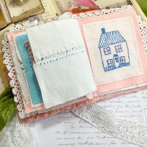 May include: A handmade fabric journal with a pink cover and a white fabric page with an embroidered blue and pink house. The journal is decorated with lace and is surrounded by vintage papers.