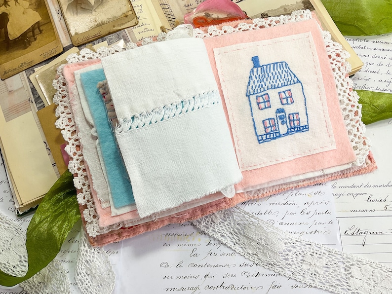 May include: A handmade fabric journal with a pink cover and a white fabric page with an embroidered blue and pink house. The journal is decorated with lace and is surrounded by vintage papers.
