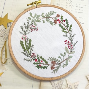 May include: Embroidered wreath on a white fabric, set in a wooden hoop. The wreath features green leaves, red berries, and brown pinecones. A festive, handcrafted piece for home decor.