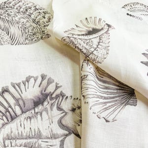 May include: Cream-coloured fabric with a repeating pattern of detailed seashell illustrations in grey and black. The design includes various shell shapes and textures, creating a coastal or nautical theme. The fabric is folded, showing the pattern's continuity.