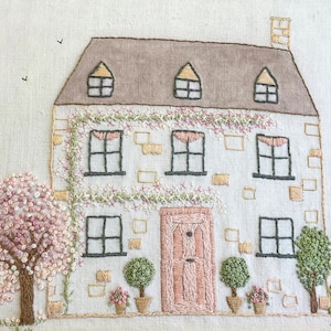 Può includere: Opera di ricamo raffigurante un cottage bianco con una porta rosa e un albero fiorito rosa. Il cottage ha un tetto marrone e finestre con cornici nere. Lo sfondo è un tessuto beige chiaro.