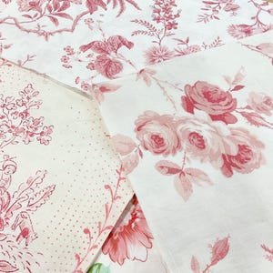 Könnte beinhalten: Nahaufnahme von mehreren weißen Stoffstücken mit rosa Blumenmustern. Ein Stück zeigt große Rosen, ein anderes ein Toile-de-Jouy-Design mit Figuren und andere botanische und gepunktete Muster. Die Stoffe sind wahrscheinlich für Bastelarbeiten oder Heimdekoration gedacht.