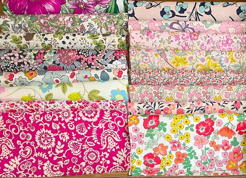 May include: A selection of floral fabric bundles in a range of colours. The fabrics display diverse floral patterns, including pink, white, green, and yellow flowers. The bundles are neatly stacked, showcasing the different designs.