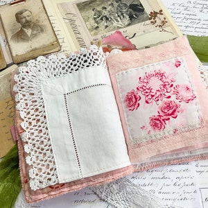 May include: A pink fabric journal with a white lace trim and a floral fabric insert. The journal is open to reveal a white page with a pink floral pattern.