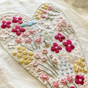 May include: A heart-shaped embroidery design featuring a variety of flowers in shades of pink, red, and yellow. The flowers are embroidered on a white linen fabric.