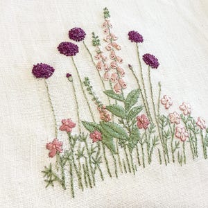 Könnte beinhalten: Gesticktes Blumendesign mit lila, rosa und grünen Blumen auf cremefarbenem Stoff. Die Stickerei zeigt detaillierte Stiche verschiedener Blumenarten, Blätter und Stiele und erzeugt ein strukturiertes, handwerkliches Aussehen. Geeignet für Heimdekoration oder Textilkunst.