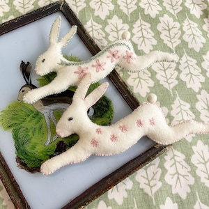 May include: Two white felt rabbit ornaments with pink floral embroidery and small white pom-poms for tails. The rabbits are posed in a running position. They are displayed on a framed picture with a green and white leaf pattern background.