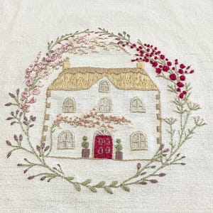 Op de afbeelding: Een borduurwerk op gebroken wit textiel met een huisje met een rieten dak en een felrode deur. Het huis is omringd door een cirkelvormige krans van geborduurde bloemen in tinten roze, donkerrood en groen blad met kleine bloemknoppen.