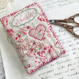 May include: A rectangular fabric needle case featuring cream-colored quilted material with hand-stitched red and pink floral embroidery, a central heart motif, and an oval patch with the word "Needles" embroidered in pink thread on the front.