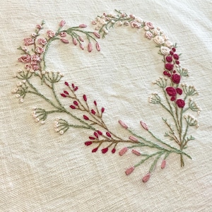 May include: A heart-shaped wreath embroidered on a white linen fabric. The wreath is made of green leaves, pink and white flowers, and red berries.