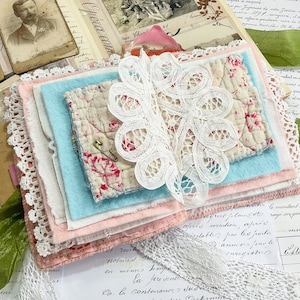 May include: A stack of fabric scraps in various shades of pink, blue, and white, with a delicate white lace overlay. The fabric scraps are arranged in a layered fashion, creating a visually appealing and textured composition.