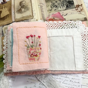 May include: A pink fabric book with a white lace trim. The front cover features an embroidered image of a potted plant with red flowers. The book is open to reveal a blank page.