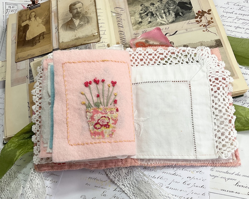 May include: A pink fabric book with a white lace trim. The front cover features an embroidered image of a potted plant with red flowers. The book is open to reveal a blank page.