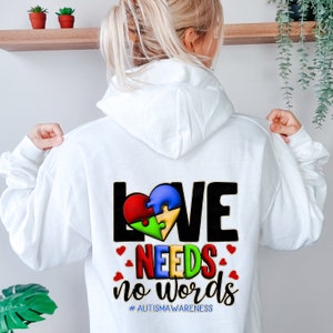 May include: White hoodie with a black and gold graphic on the back. The graphic features a puzzle piece heart with the words "LOVE NEEDS NO WORDS" and the hashtag "#AUTISMAWARENESS".