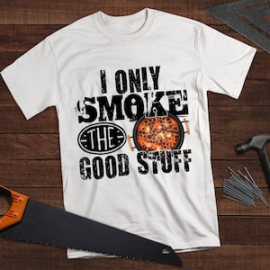 May include: White t-shirt with black text that reads "I only smoke the good stuff". The text is distressed and has a vintage look. There is a black and white illustration of a grill with glowing coals.