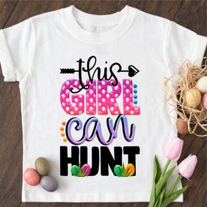 May include: White t-shirt with a pink, purple, and blue graphic that says "This Girl Can Hunt". The graphic has polka dots and a small arrow with a heart.