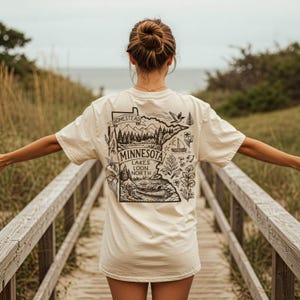 May include: Cream-colored t-shirt featuring a black and white graphic of the Minnesota state outline with the text "MINNESOTA LAKES LOON NORTH". The design includes illustrations of trees, mountains, a sailboat, and a loon.