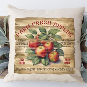 May include: A square, off-white pillow with a printed design of red apples and green leaves on a wooden background. The text "FARM FRESH APPLES" is arched across the top. Additional text includes "CRISP & FRESH", "PICK YOUR OWN", ".25¢ EACH", and apple varieties.