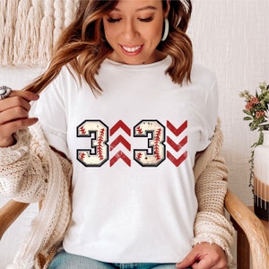 May include: White t-shirt with the number 333 printed in red and white. The numbers are made to look like baseballs with red stitching. The numbers are flanked by red and white chevron patterns.