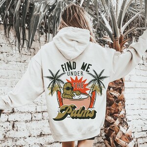 May include: White hoodie with a graphic print of a cartoon cactus wearing sunglasses and relaxing in a coconut. The text "Find Me Under The Palms" is printed on the hoodie.