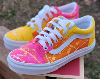 May include: A pair of custom-painted Vans Old Skool sneakers with a pink, orange, and yellow splatter paint design. The shoes are sitting on top of a red Vans shoebox.