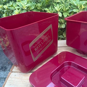May include: Red plastic ice bucket with the text "Johnnie Walker RED LABEL" on a gold label. The bucket is rectangular with a square lid. The background is a wooden surface and green foliage.