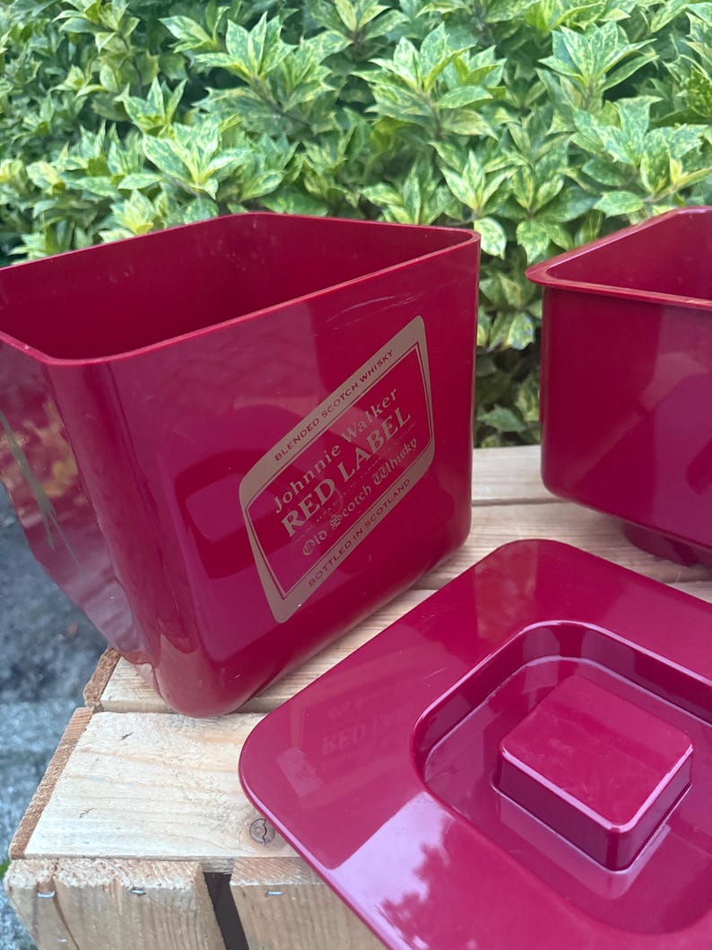May include: Red plastic ice bucket with the text "Johnnie Walker RED LABEL" on a gold label. The bucket is rectangular with a square lid. The background is a wooden surface and green foliage.