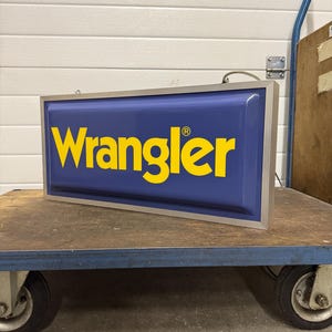 May include: A rectangular sign with the word "Wrangler" in yellow letters on a blue background. The sign has a silver frame and is sitting on a blue cart with black wheels.