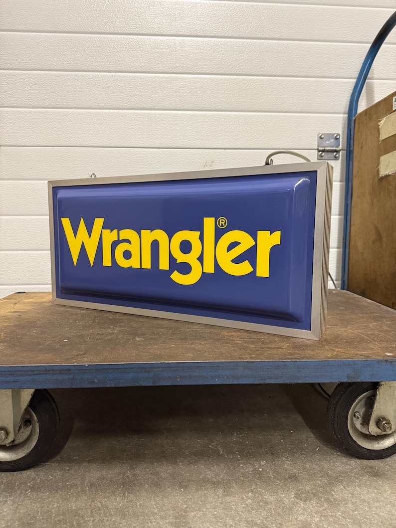 May include: A rectangular sign with the word "Wrangler" in yellow letters on a blue background. The sign has a silver frame and is sitting on a blue cart with black wheels.