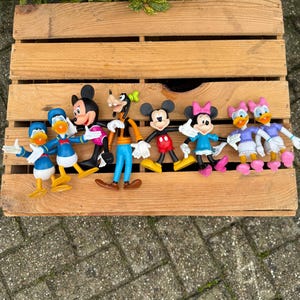 Könnte beinhalten: Eine Sammlung von Disney-Charakterfiguren, darunter Micky Maus, Minnie Maus, Donald Duck, Daisy Duck und Goofy, ist auf einer Holzkiste angeordnet. Die Figuren sind farbenfroh und weisen klassische Cartoon-Designs auf. Die Kiste ist aus hellbraunem Holz.