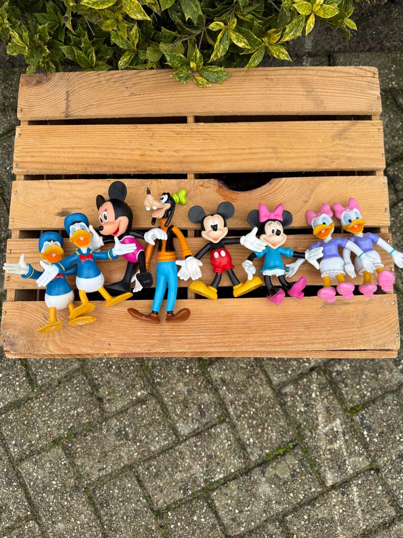 Pode incluir: Uma cole&ccedil;&atilde;o de figuras de personagens da Disney, incluindo Mickey Mouse, Minnie Mouse, Pato Donald, Margarida e Pateta, est&aacute; disposta em uma caixa de madeira. As figuras s&atilde;o coloridas e apresentam designs cl&aacute;ssicos de desenhos animados. A caixa &eacute; de madeira marrom claro.
