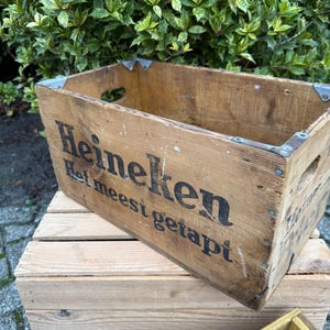 May include: A vintage wooden crate, branded with the Heineken logo and text in black. The crate features metal corner reinforcements and hand-holds. It rests on another wooden crate. Green foliage is visible in the background.