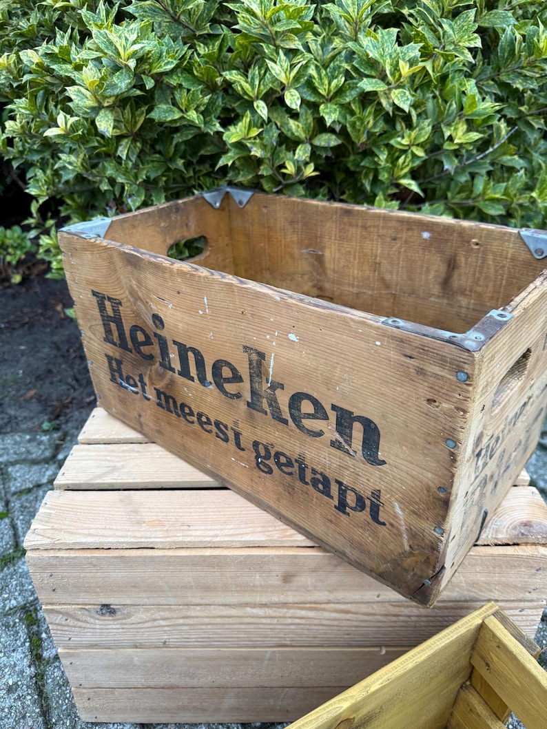May include: A vintage wooden crate, branded with the Heineken logo and text in black. The crate features metal corner reinforcements and hand-holds. It rests on another wooden crate. Green foliage is visible in the background.