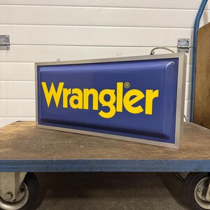 May include: A rectangular sign with the word "Wrangler" in yellow letters on a blue background. The sign has a silver frame and is sitting on a blue cart with wheels. The sign is lit.