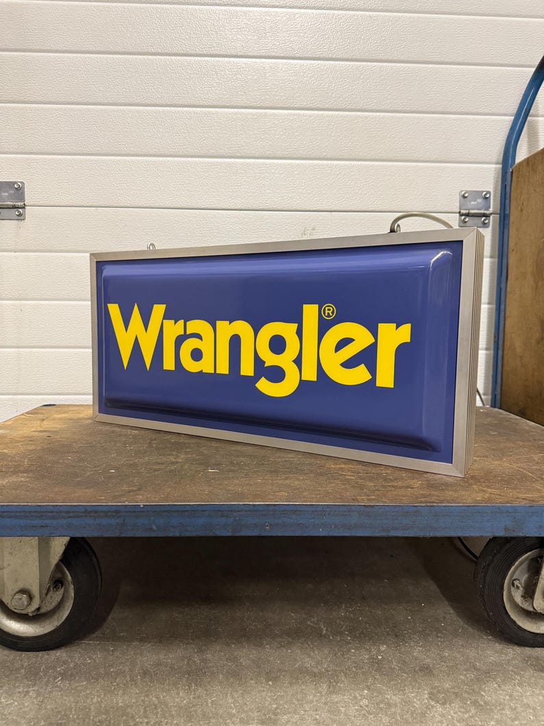 May include: A rectangular sign with the word "Wrangler" in yellow letters on a blue background. The sign has a silver frame and is sitting on a blue cart with wheels. The sign is lit.