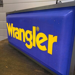 May include: A rectangular sign with the word "Wrangler" in yellow letters on a blue background. The sign has a black metal frame and a curved handle at the top. The registered trademark symbol is next to the word Wrangler.