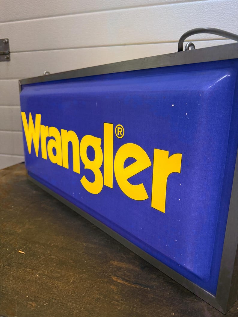 May include: A rectangular sign with the word "Wrangler" in yellow letters on a blue background. The sign has a black metal frame and a curved handle at the top. The registered trademark symbol is next to the word Wrangler.