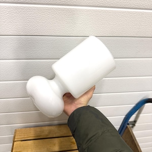 May include: A white, opaque glass vase with a bulbous base and a cylindrical top. The vase is held at an angle, showcasing its unique shape and design. The background is a white, textured surface.